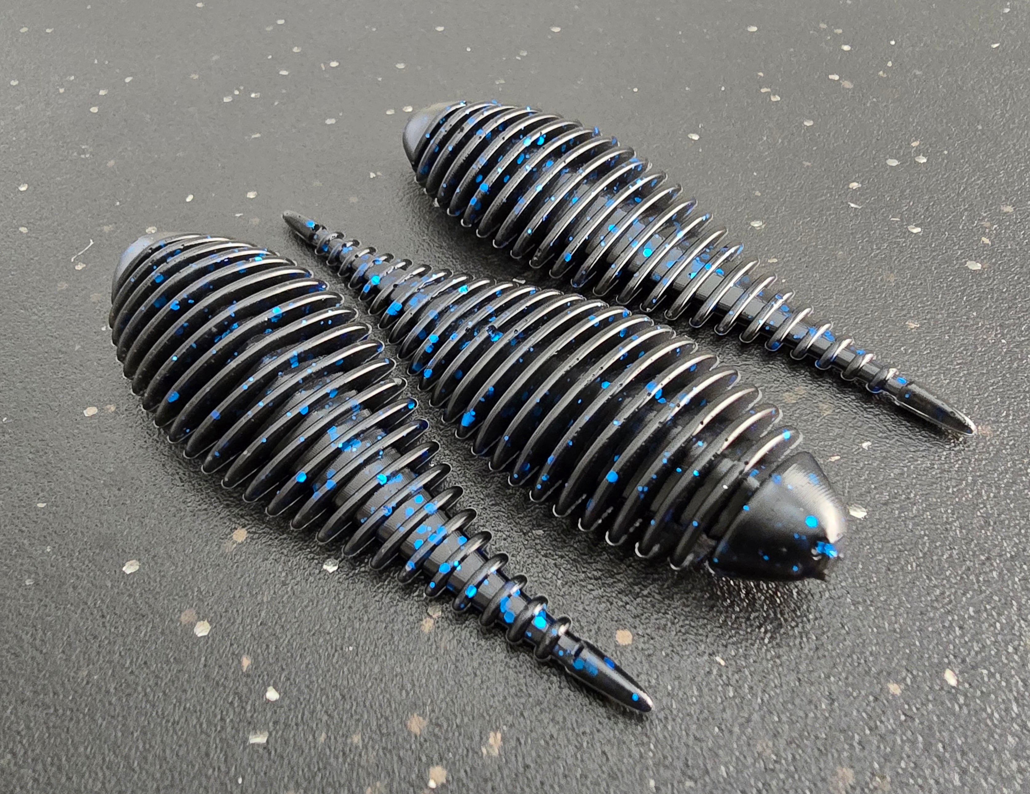 Three black and blue Ambush Gilly fishing soft plastic baits on a gray surface